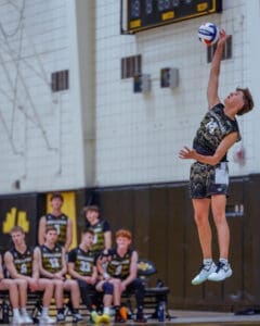 High school volleyball player jumps to serve during a match, with teammates watching in the background.