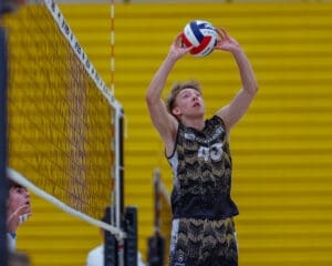 High school volleyball player sets the ball during match, focused on gameplay with teammate in the background.