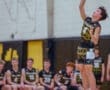 A volleyball player jumps to serve, with teammates watching in the background during a competitive match.