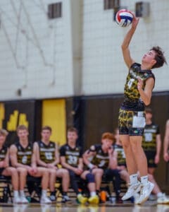 A volleyball player jumps to serve, with teammates watching in the background during a competitive match.