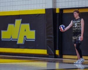 High school volleyball player in action, ready to serve with a ball in hand against a bold yellow and black background.