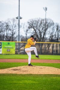 A baseball pitcher in a yellow jersey winds up on the mound during a game, with a blurred background of trees.