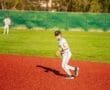 A baseball player fields the ball on a vibrant red infield during an evening practice under the sun.