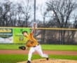 A young baseball pitcher in an orange jersey throws a pitch from the mound during a game on a grassy field.