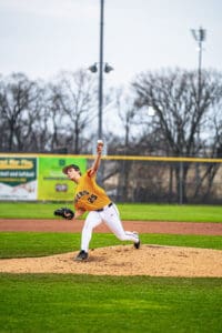 A young baseball pitcher in an orange jersey throws a pitch from the mound during a game on a grassy field.