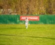 A baseball player in a white uniform walks in the outfield near an Armstrong sign and green fence under a clear sky.
