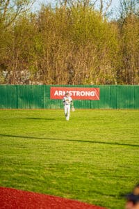 A baseball player in a white uniform walks in the outfield near an Armstrong sign and green fence under a clear sky.