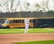 High school baseball player stands on the field, ready for action, with a school bus parked in the background.