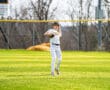 Young baseball player wearing a striped uniform prepares to throw on a sunny field with a scoreboard in the background.