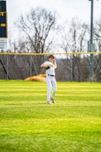Young baseball player wearing a striped uniform prepares to throw on a sunny field with a scoreboard in the background.