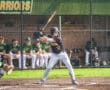Baseball player at bat during a game, with teammates in green uniforms visible on the bench in the background.