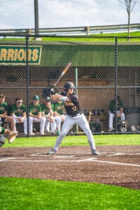 Baseball player at bat during a game, with teammates in green uniforms visible on the bench in the background.