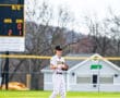Baseball player in striped uniform stands on field near scoreboard, focused on the game in a spring setting.