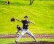 A young pitcher in a black jersey throws the ball from the mound during a baseball game on a sunny day.