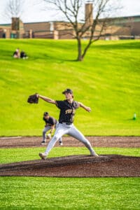 A young pitcher in a black jersey throws the ball from the mound during a baseball game on a sunny day.