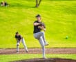 Young baseball player on the pitcher's mound prepares to throw, with teammates in the background and a vibrant green field.