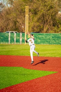 Baseball player leaps to catch a ball during the game on a vibrant red and green field under clear skies.
