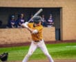 A young baseball player in an orange jersey prepares to hit, with teammates watching from the dugout in the background.
