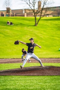 High school baseball pitcher in motion on the mound, with players and greenery in the background.