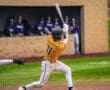 Young baseball player swings bat at home plate during a game, with teammates watching from the dugout in the background.