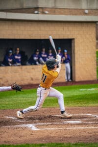Young baseball player swings bat at home plate during a game, with teammates watching from the dugout in the background.