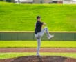 Baseball pitcher in mid-windup on the mound, wearing a black jersey and grey pants, with a vibrant green field behind.