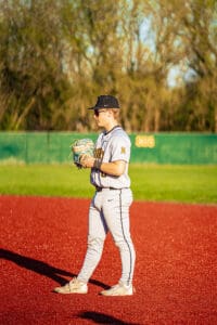 Player in a grey baseball uniform stands on a red field, preparing to pitch with glove in hand under bright sunlight.