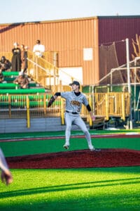 A young baseball pitcher in gray Tigers uniform winds up to throw from the mound, with spectators in the background.