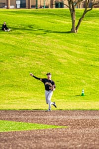 Teenage baseball player in a black jersey throwing a ball on a sunny day at a green field.