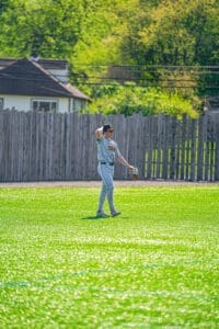 Player in gray baseball uniform stands on a sunny field, preparing for the game, glove in hand, background of trees.