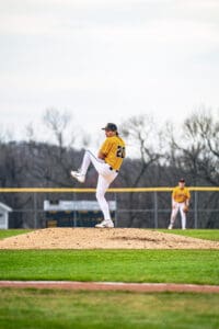 Young baseball pitcher in action on the mound during a game, with teammate in the background on the field.