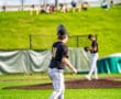 A young baseball player in black jersey stands on first base, while teammates practice on a sunny field.