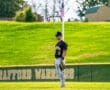 Young baseball player in a black uniform stands on a sunny field, focused on the game with an American flag in the background.