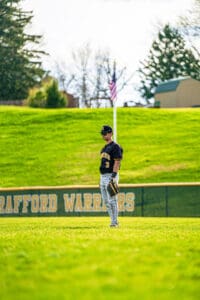 Young baseball player in a black uniform stands on a sunny field, focused on the game with an American flag in the background.