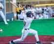 High school baseball player swings at a pitch on a colorful field while teammates cheer in the background.