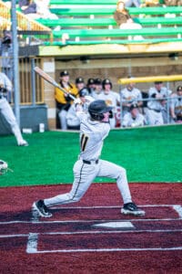 High school baseball player swings at a pitch on a colorful field while teammates cheer in the background.