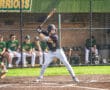 Baseball player poised to swing bat at home plate, with teammates in the dugout during a sunny game day.