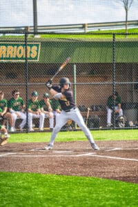 Baseball player poised to swing bat at home plate, with teammates in the dugout during a sunny game day.