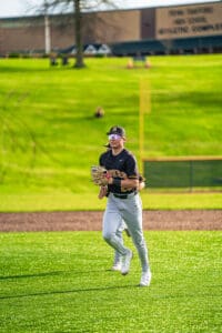High school baseball player running on the field, wearing a uniform and sunglasses at an athletic complex.