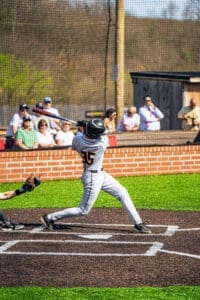 Youth baseball player swings bat, aiming to hit the ball, with spectators watching in the background on a sunny day.