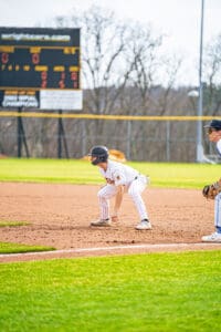 A baseball player in a striped uniform prepares to run, with a scoreboard and teammate in the background on a sunny day.
