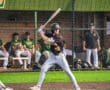 Young baseball player swings a bat at home plate with teammates in the dugout during a sunny game day.
