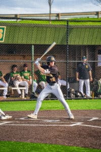 Young baseball player swings a bat at home plate with teammates in the dugout during a sunny game day.