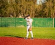 Youth baseball player stands on a red field, wearing a uniform, ready for action in a sunny park setting.