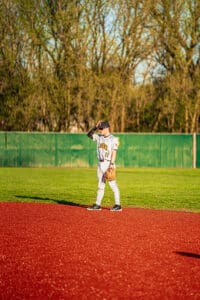 Youth baseball player stands on a red field, wearing a uniform, ready for action in a sunny park setting.