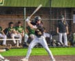 Player ready to swing a bat during a baseball game, with teammates in the dugout in the background.