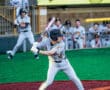 A young baseball player in a gray uniform prepares to swing a bat at home plate, with teammates in the background.