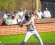 A baseball player in a gray uniform prepares to bat as spectators watch from behind the netting.