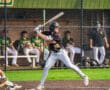 Young baseball player in black jersey swings bat on the field, with teammates on the bench in the background.