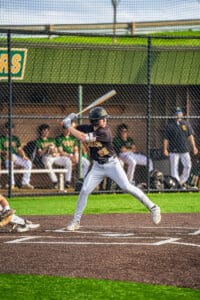 Young baseball player in black jersey swings bat on the field, with teammates on the bench in the background.